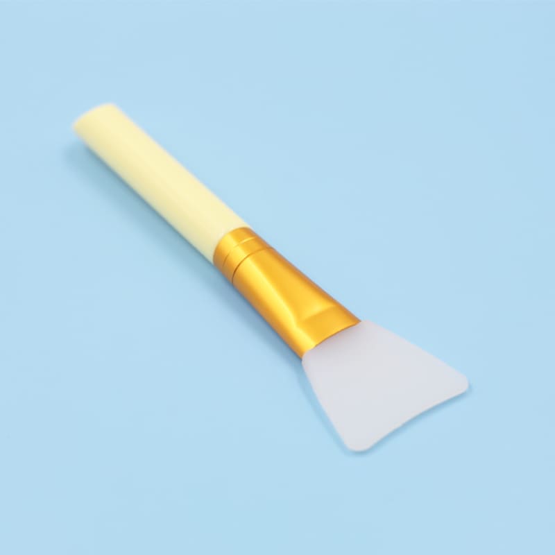 Silicone facial mask applicator brush with yellow handle and gold ferrule on a blue background.