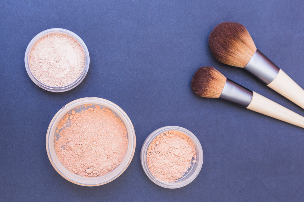 Makeup still life: Three containers of powder foundation and two makeup brushes on a dark blue background.