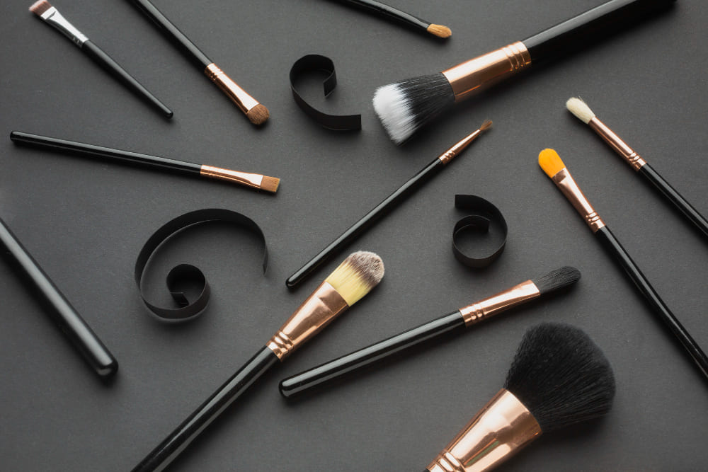 Assorted makeup brushes with black handles and copper fittings arranged with black paper curls on a dark background.