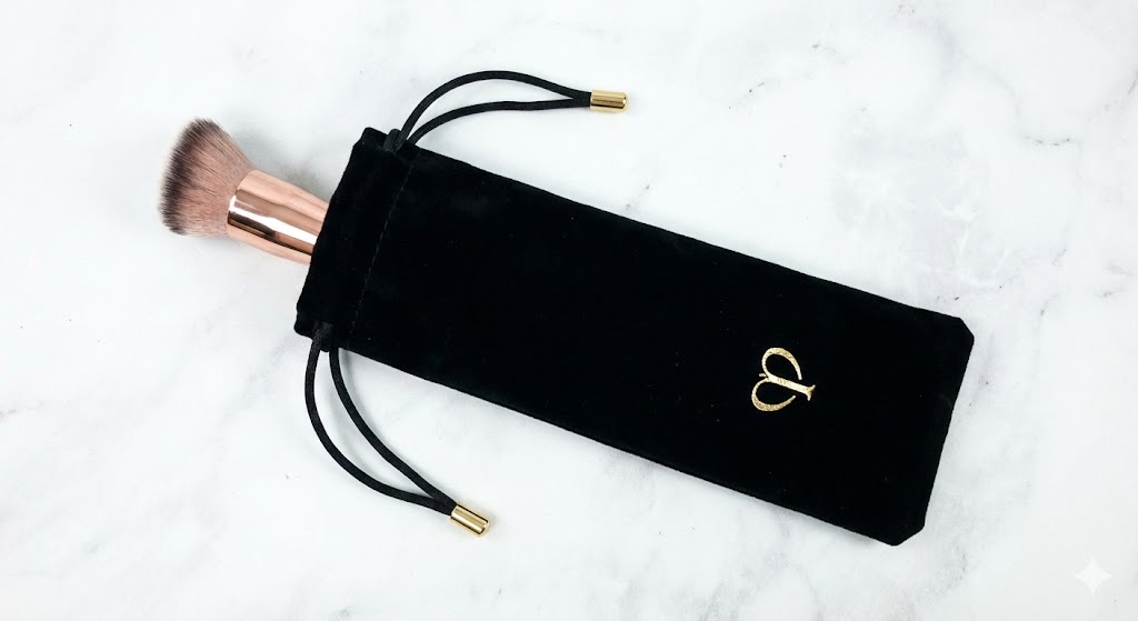 Pinceau de maquillage partiellement rangé dans une pochette à cordon en velours noir avec un logo doré sur une surface en marbre.