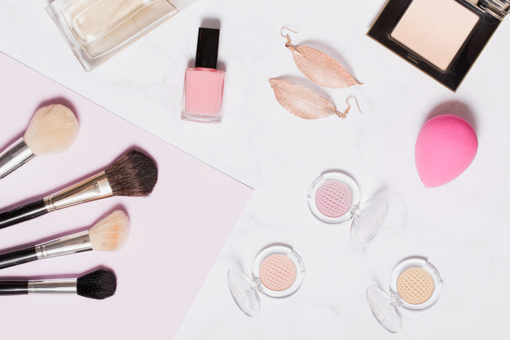 Flat lay of makeup brushes, blush, pink nail polish, perfume, leaf earrings, pink sponge, and powder on a marble and pink background.