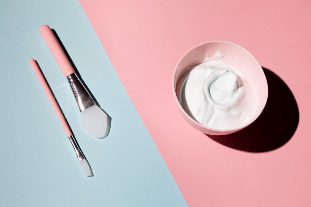 Facial mask preparation: a bowl of white cream next to two application brushes on a split pink and blue background.
