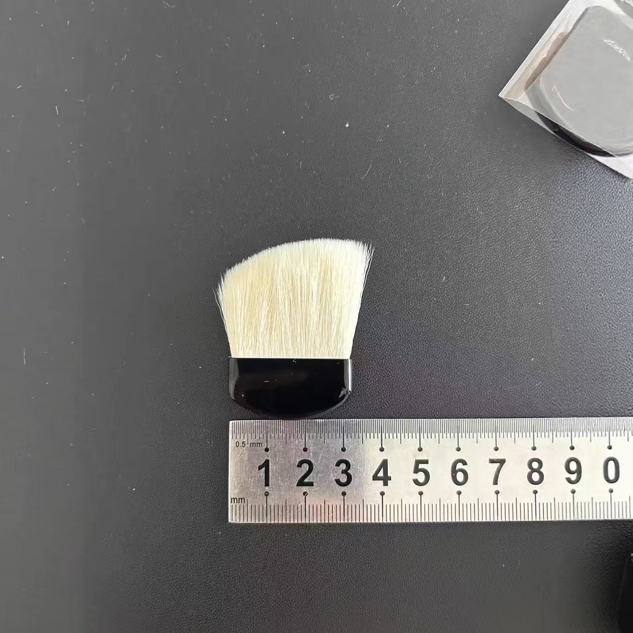 Cosmetic brush lying next to a ruler on a black surface.